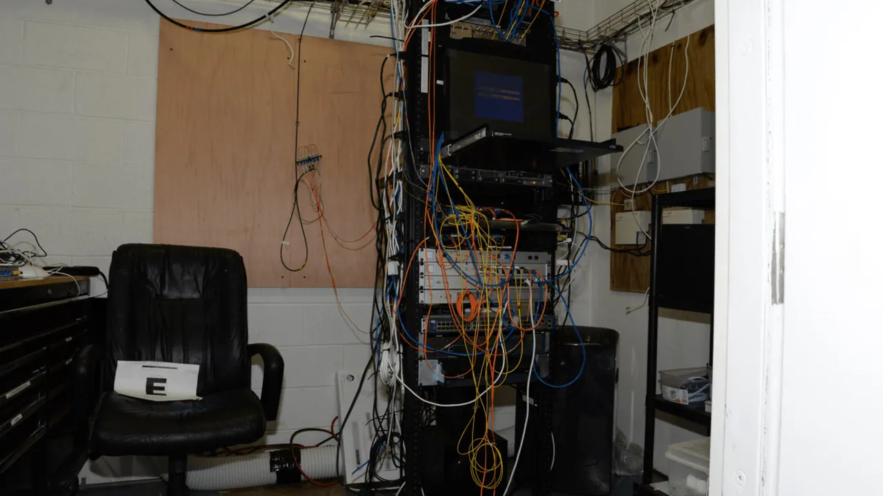 EFTA00002517 - Cluttered tech room with a server rack tangled cables and a black office chair holding a paper with E on
