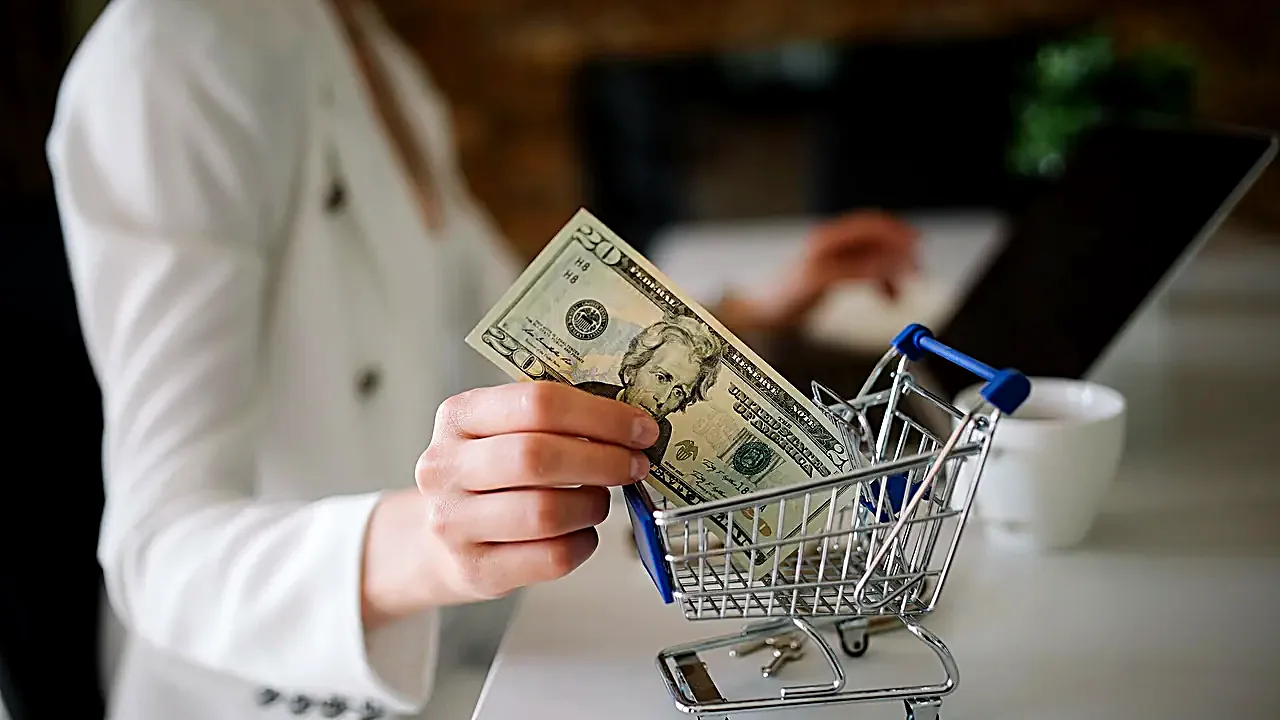 Woman shops online with a small shopping cart and cash in a modern office setting during the afternoon