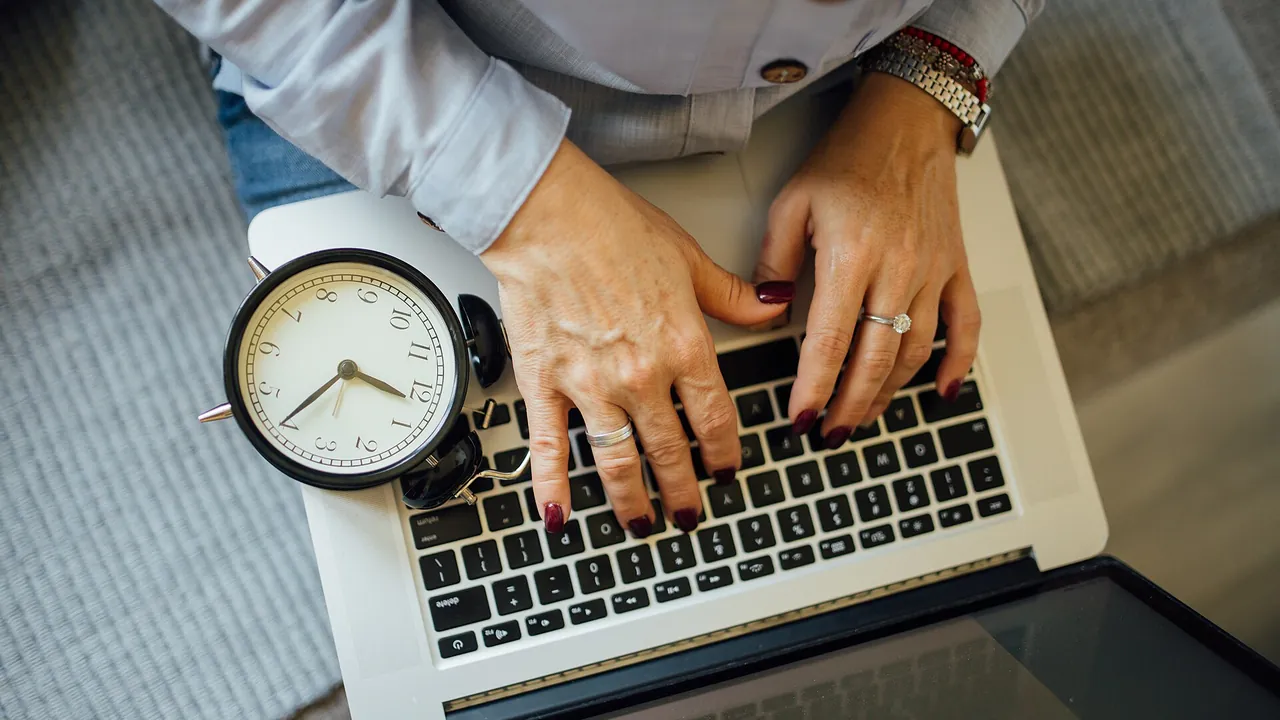 Hands are typing on a laptop keyboard while a clock rests next to the device