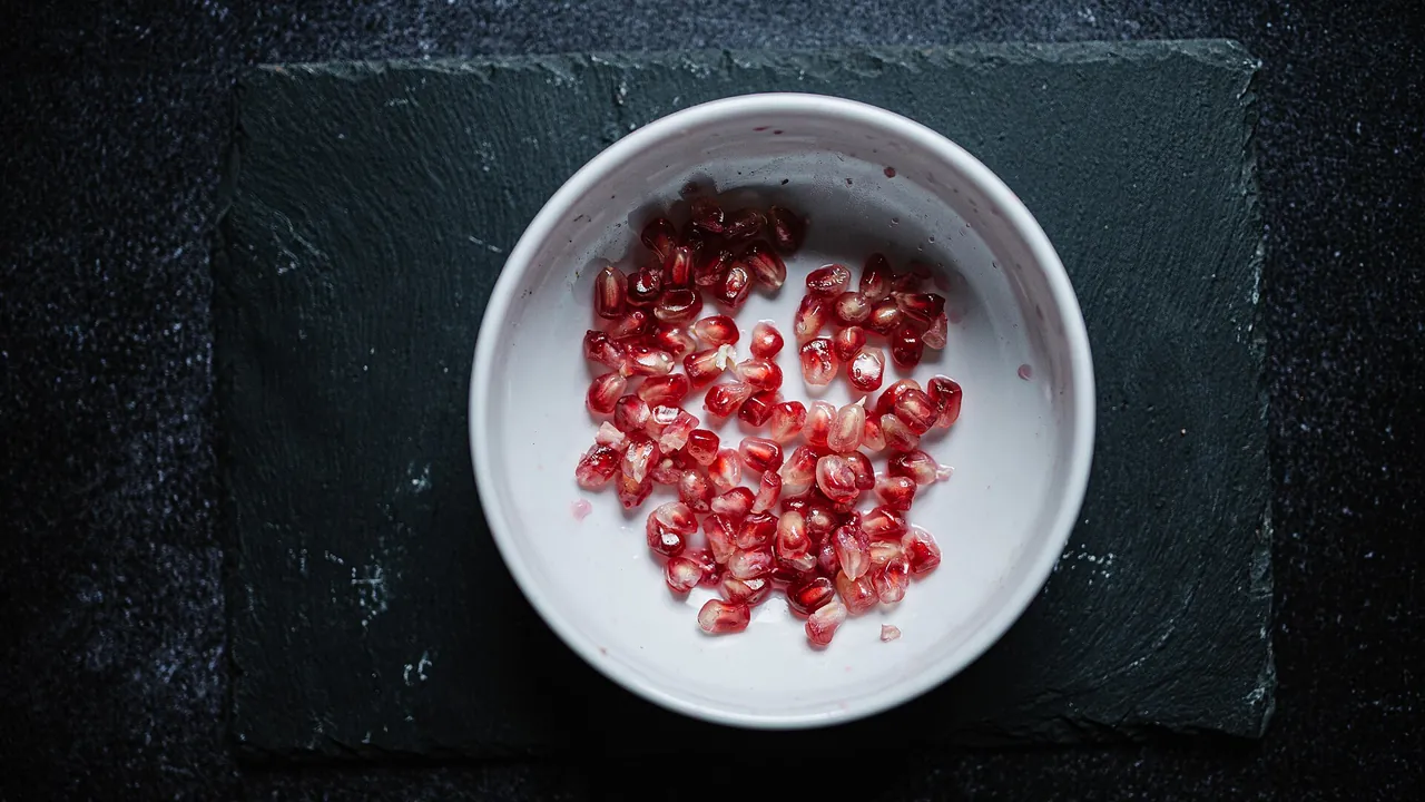 Bright red pomegranate seeds arranged in a white bowl on a dark surface suitable for culinary presentation