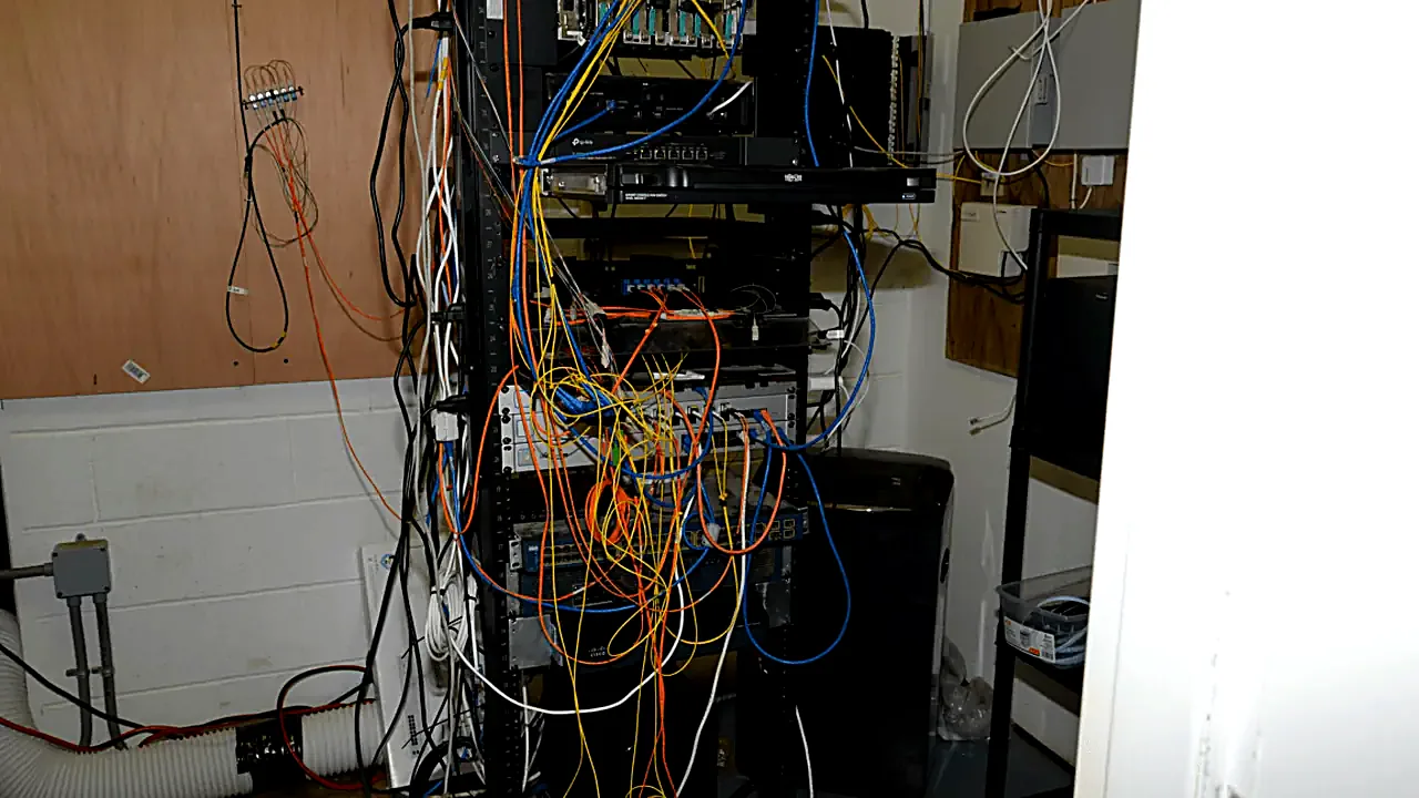 EFTA00002627 - Cluttered server rack filled with tangled cables and networking equipment in a basement room