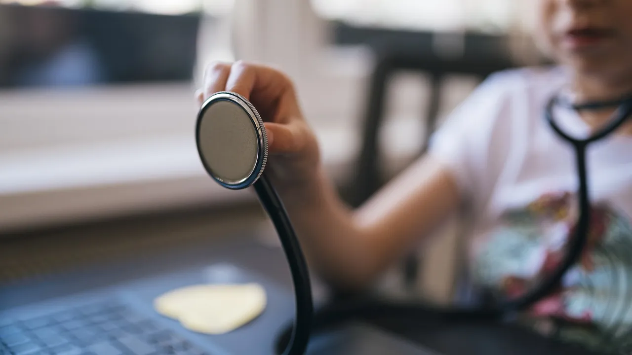 Child plays doctor with stethoscope next to laptop while engaged in imaginative pretend play at home