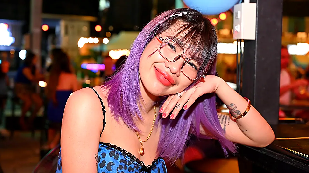 DSC 7504 Young woman with purple hair and glasses smiles warmly at the camera while resting her head on her hand at a li